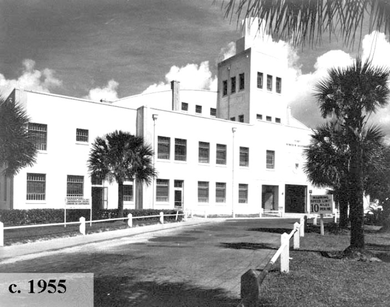 Historic black and white photo of a building from 1955 with palm trees and a sign on the roadside.