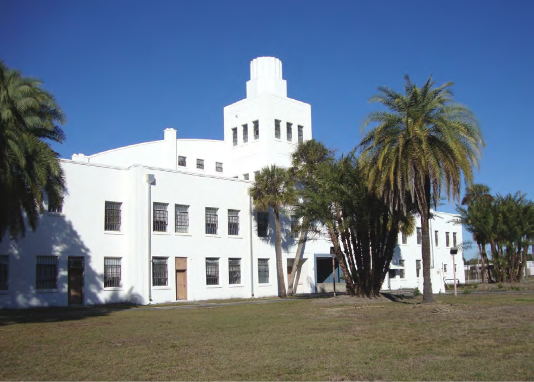 A white, art deco style building surrounded by palm trees under a clear blue sky.