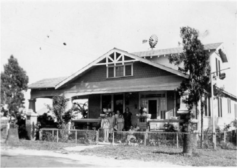 Black and white photo of a front porch with four people standing in front of a large house, surrounded by trees and a fence.