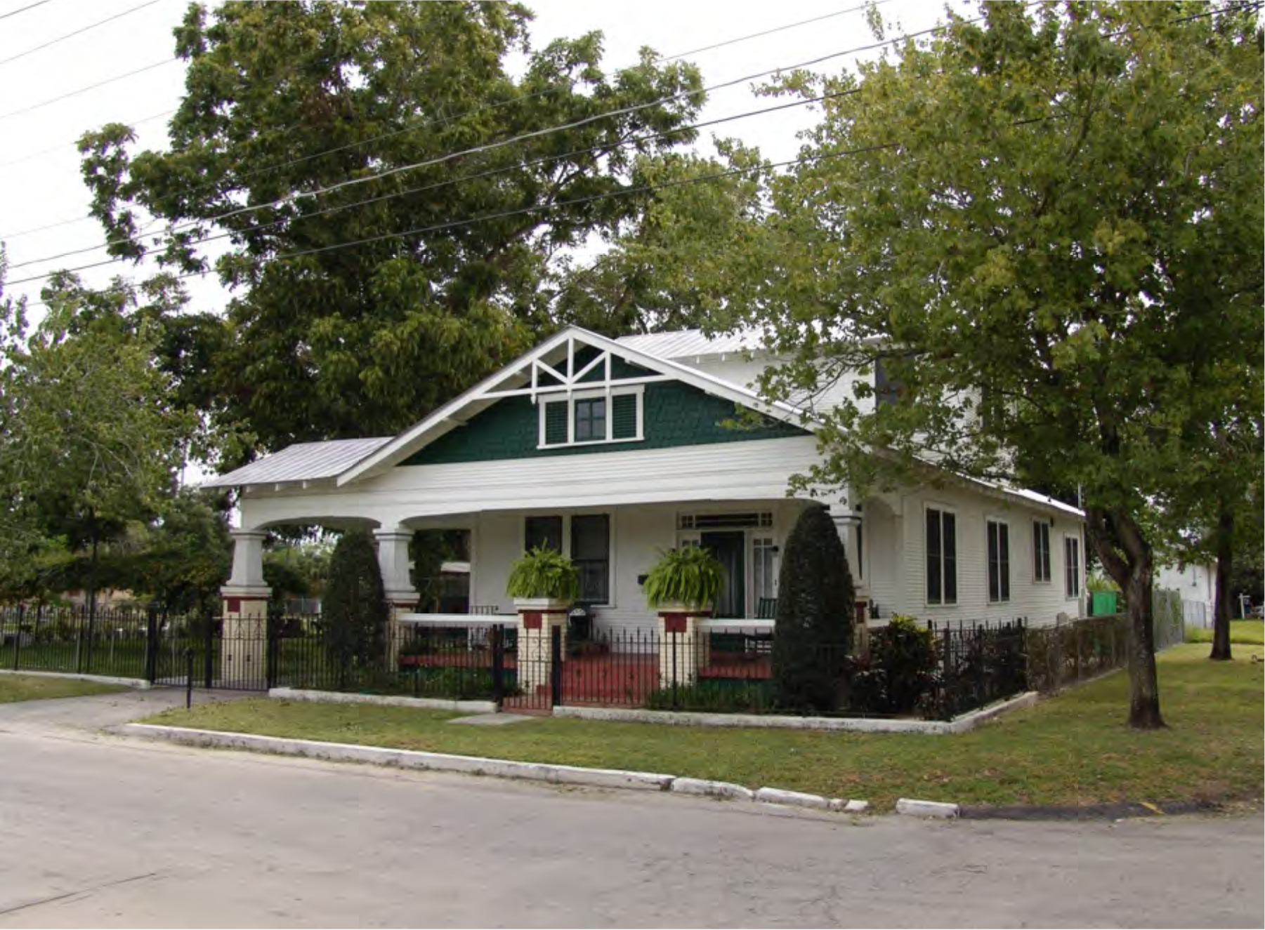 A white house with green accents and a red porch, surrounded by trees and shrubs, located on a quiet street.