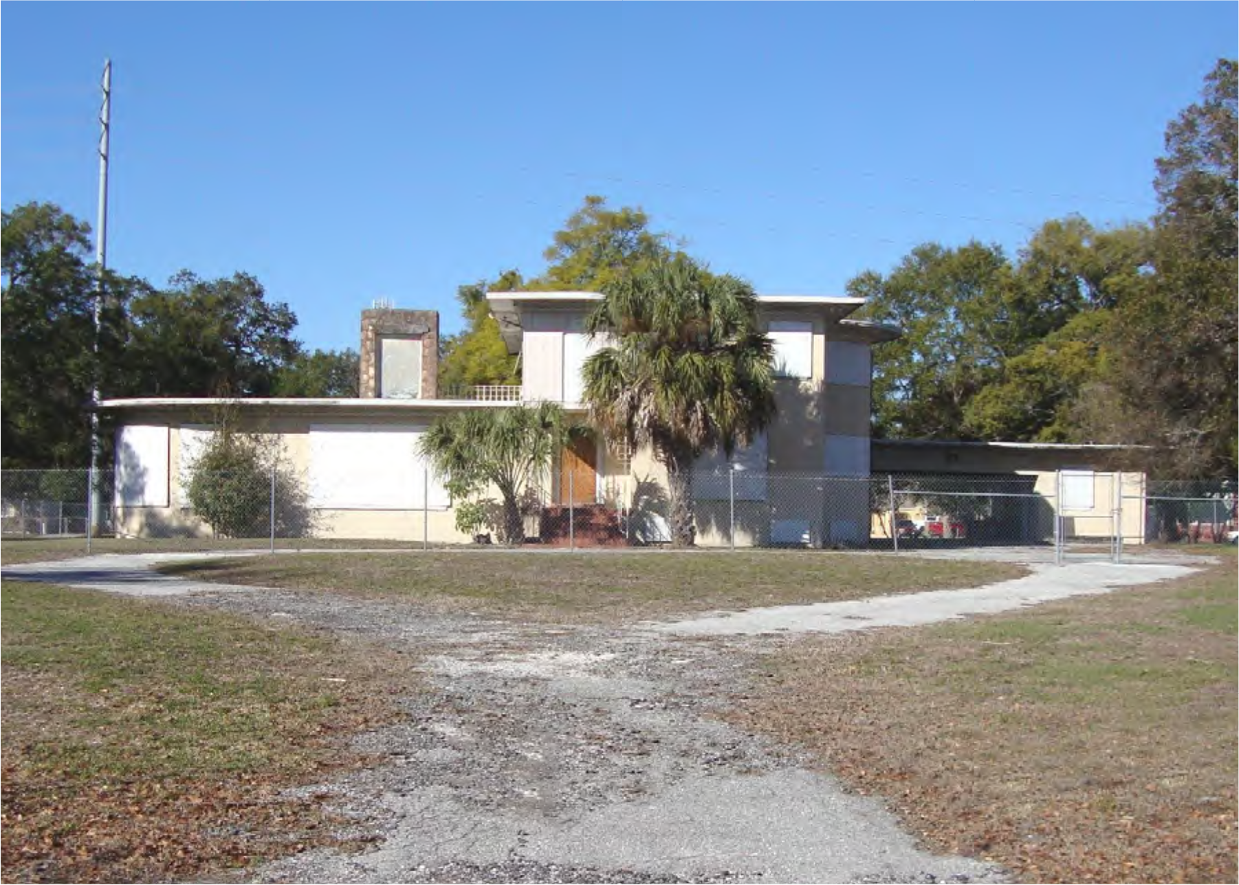 Abandoned building surrounded by a fence, featuring palm trees and a clear blue sky.