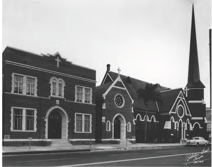 Historic brick church buildings with a tall spire, cross, and clock tower, set against a street backdrop.