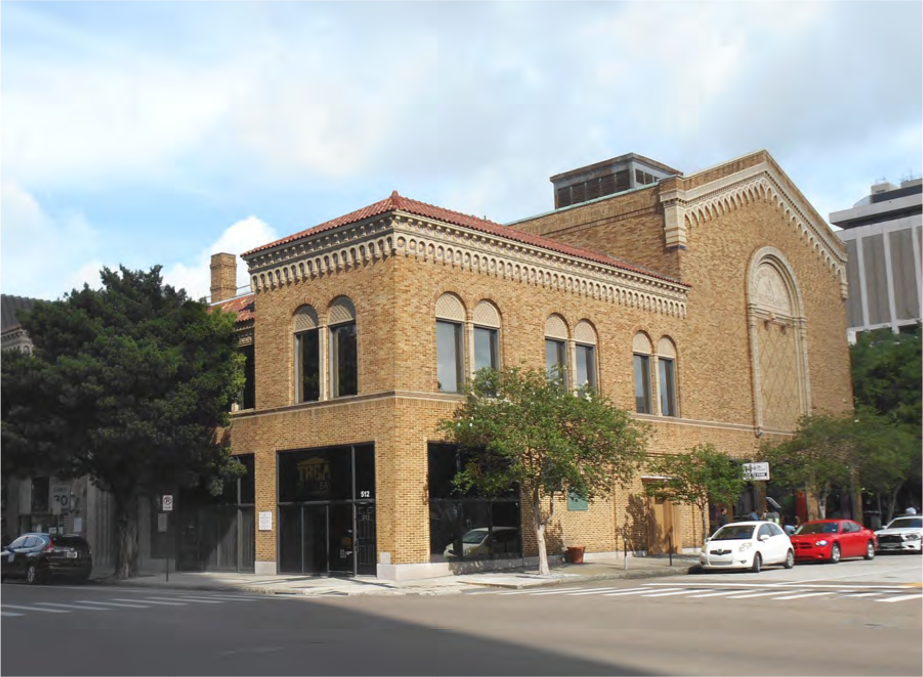 Historic brick building with arched windows and decorative cornice on a street corner, featuring trees and parked cars around it.