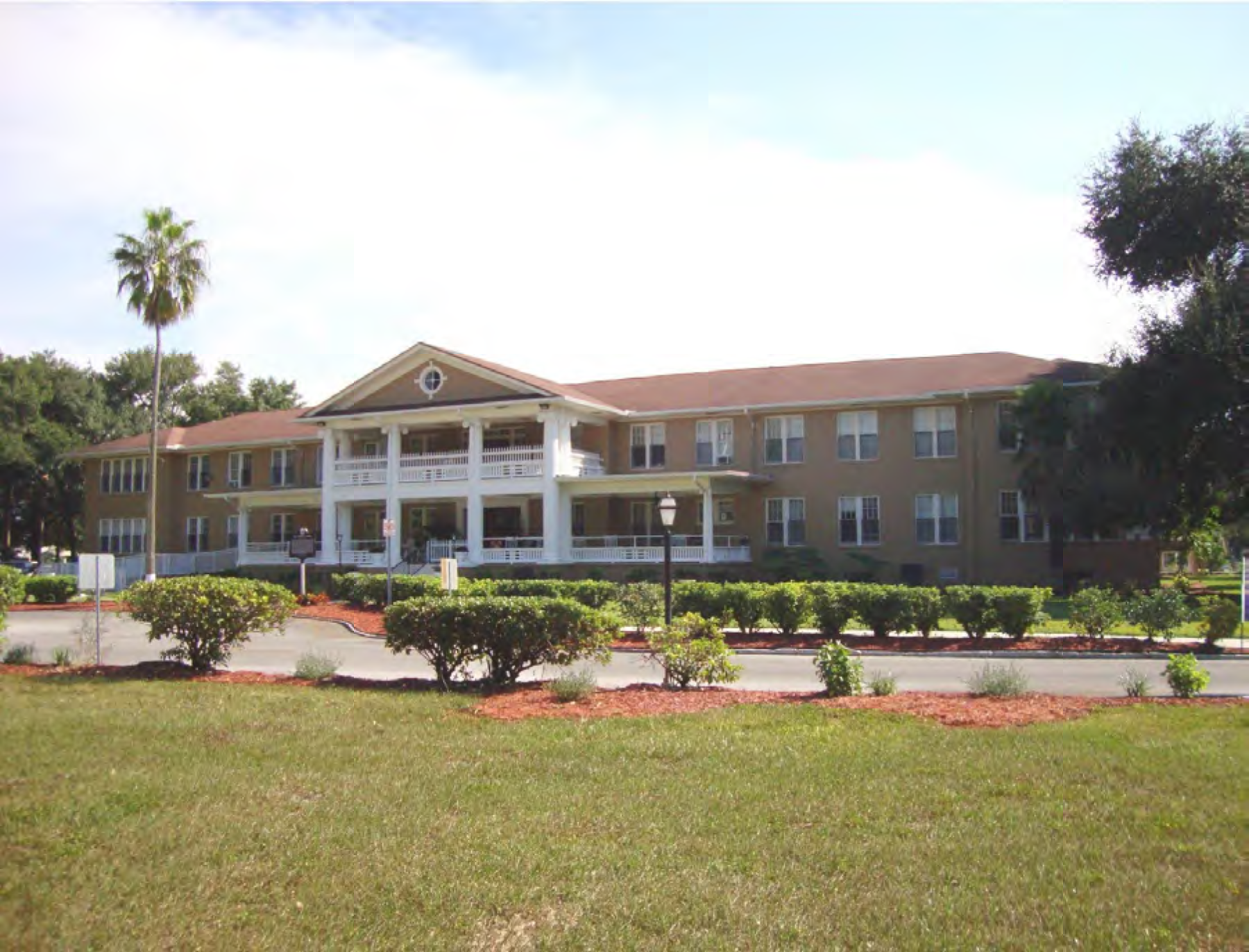A large, two-story building with a porch, surrounded by green grass and shrubs under a clear sky.