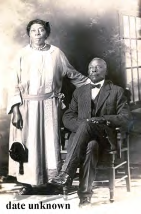 A historical black-and-white photo of a man seated and a woman standing beside him, both in vintage attire.