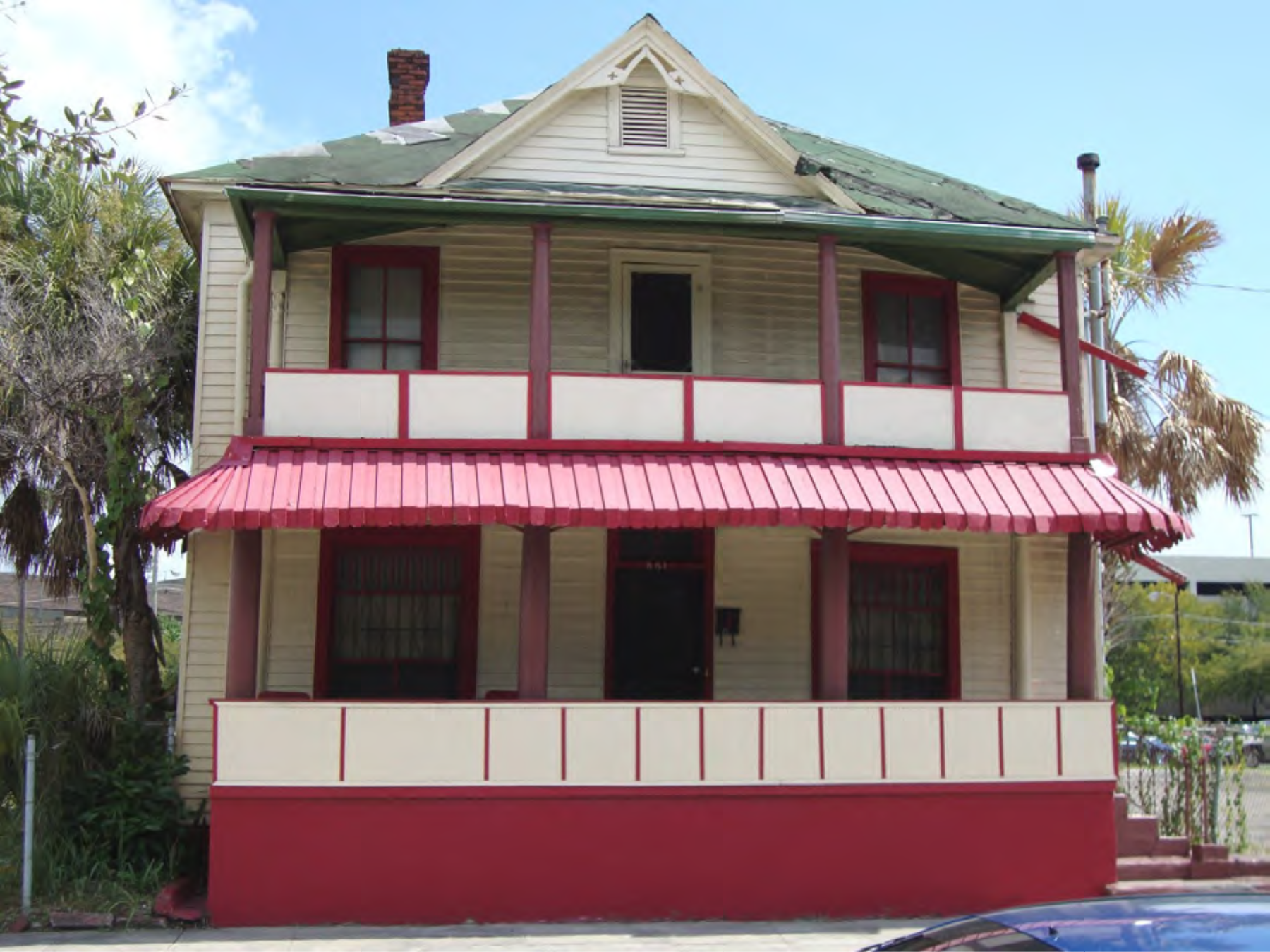 Historic two-story house with a red metal roof, white and red accents, and porch, set against a clear blue sky.