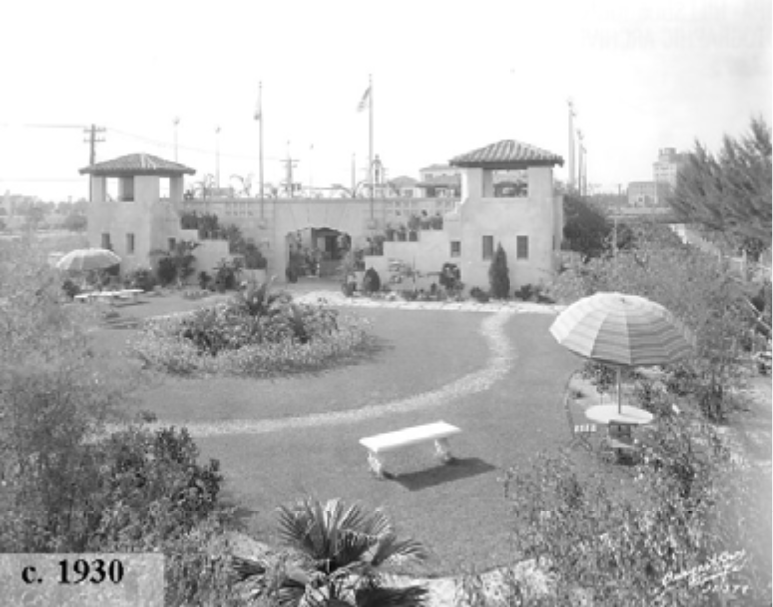 Historical black and white photo of a garden with a stone structure, benches, and umbrellas, taken around 1930.