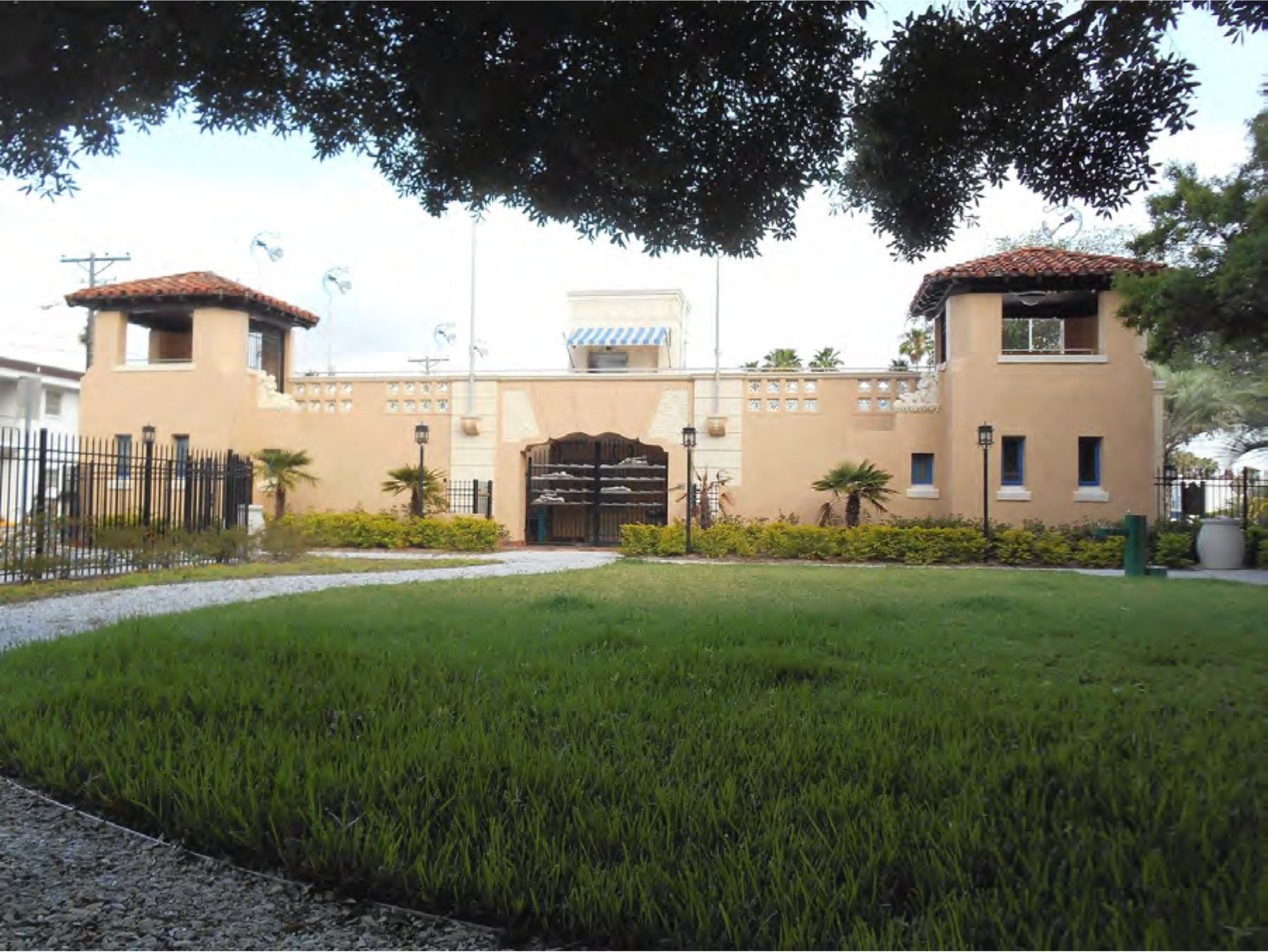 A two-story stucco building with turret-like features, surrounded by a grassy yard and decorative fencing in a sunny setting.