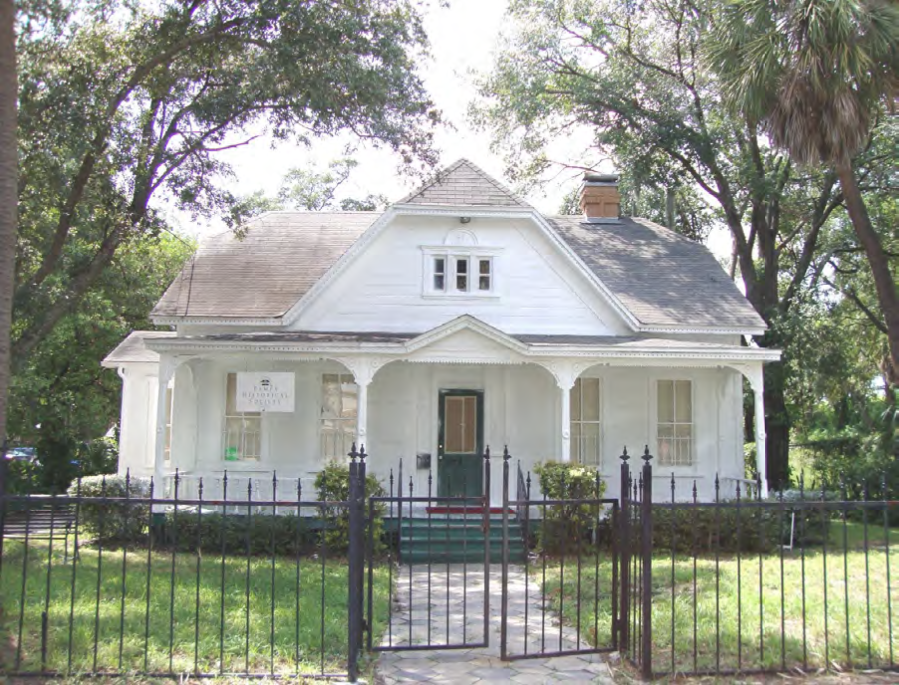 A white, historic house with a porch, surrounded by greenery and a black fence.