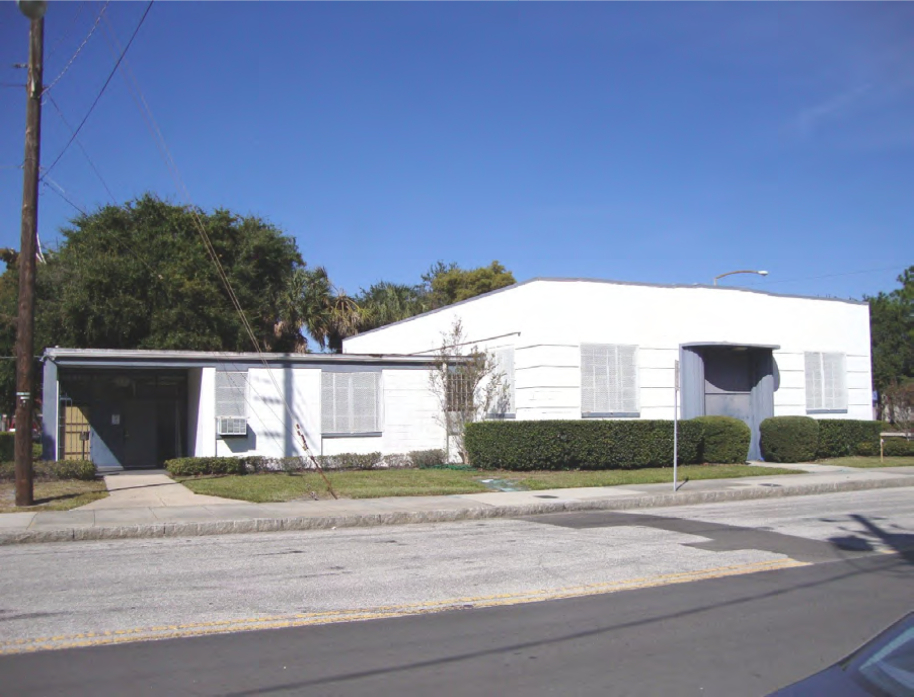 A white, modern building with windows and greenery, situated on a street under a clear blue sky.