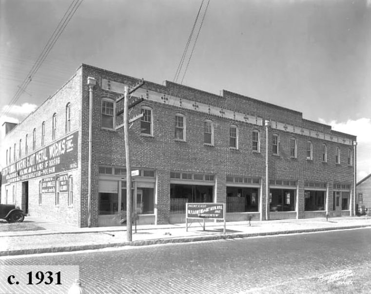 Historic black-and-white photo of a brick commercial building from 1931, featuring storefronts and vintage signage.