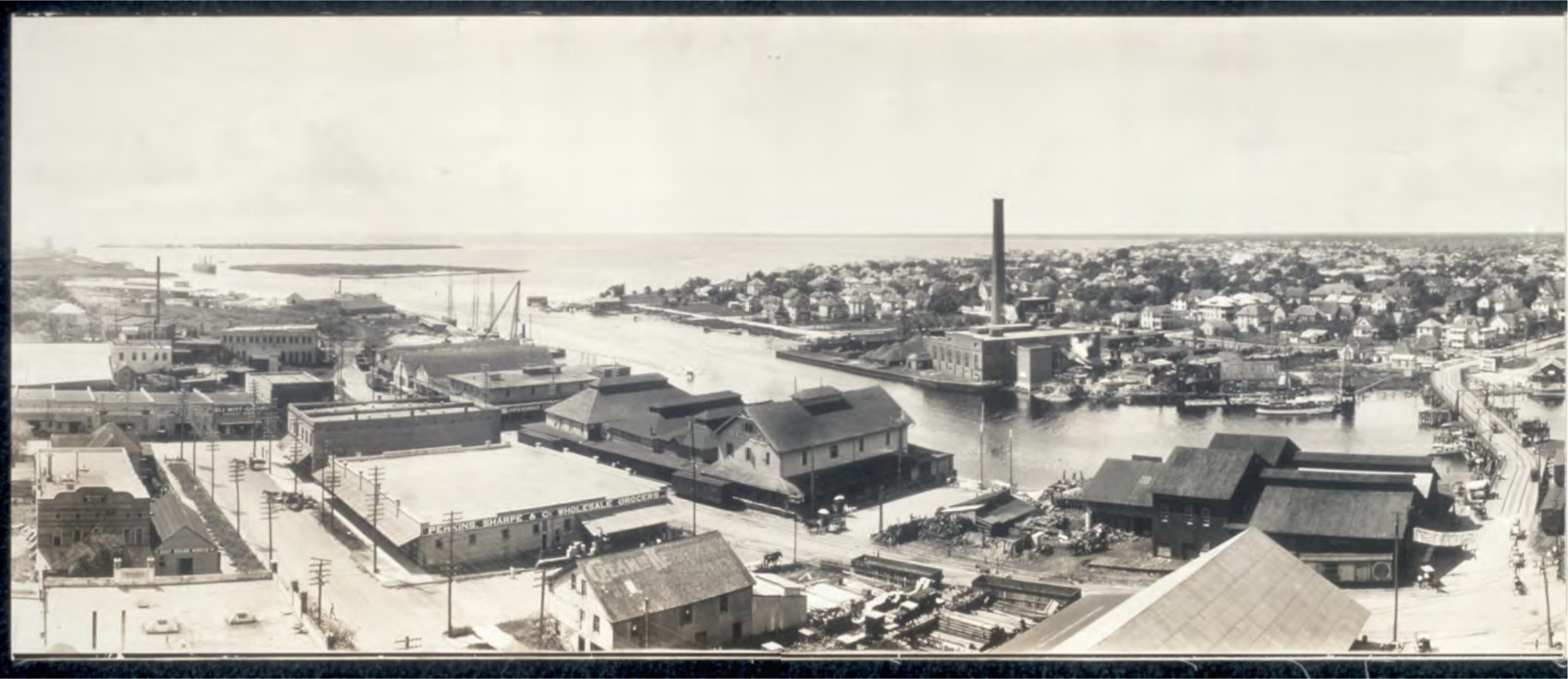 Black and white panoramic view of a coastal town with docks, warehouses, and houses by the water.