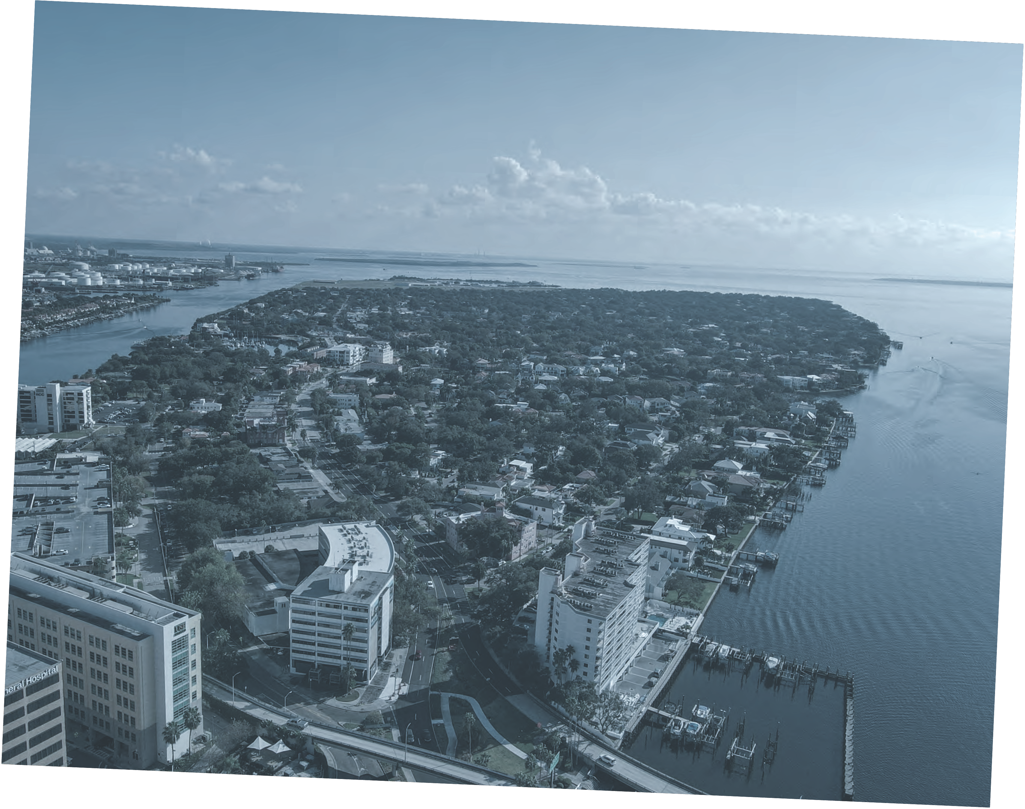 Aerial view of a waterfront cityscape featuring residential areas, greenery, and a river, under a cloudy sky.