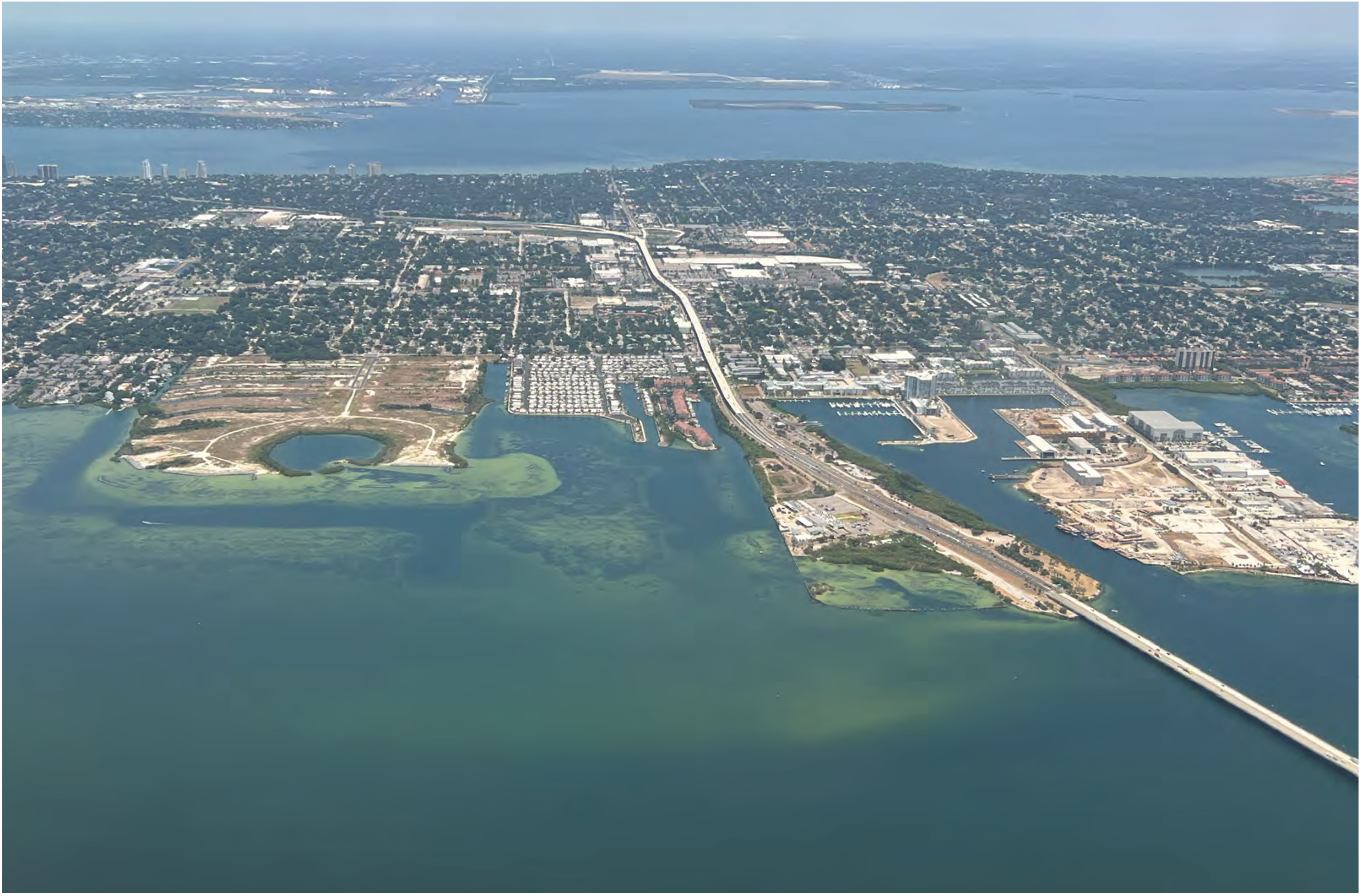 Aerial view of a coastal city with marinas, industrial areas, and green spaces along the water.