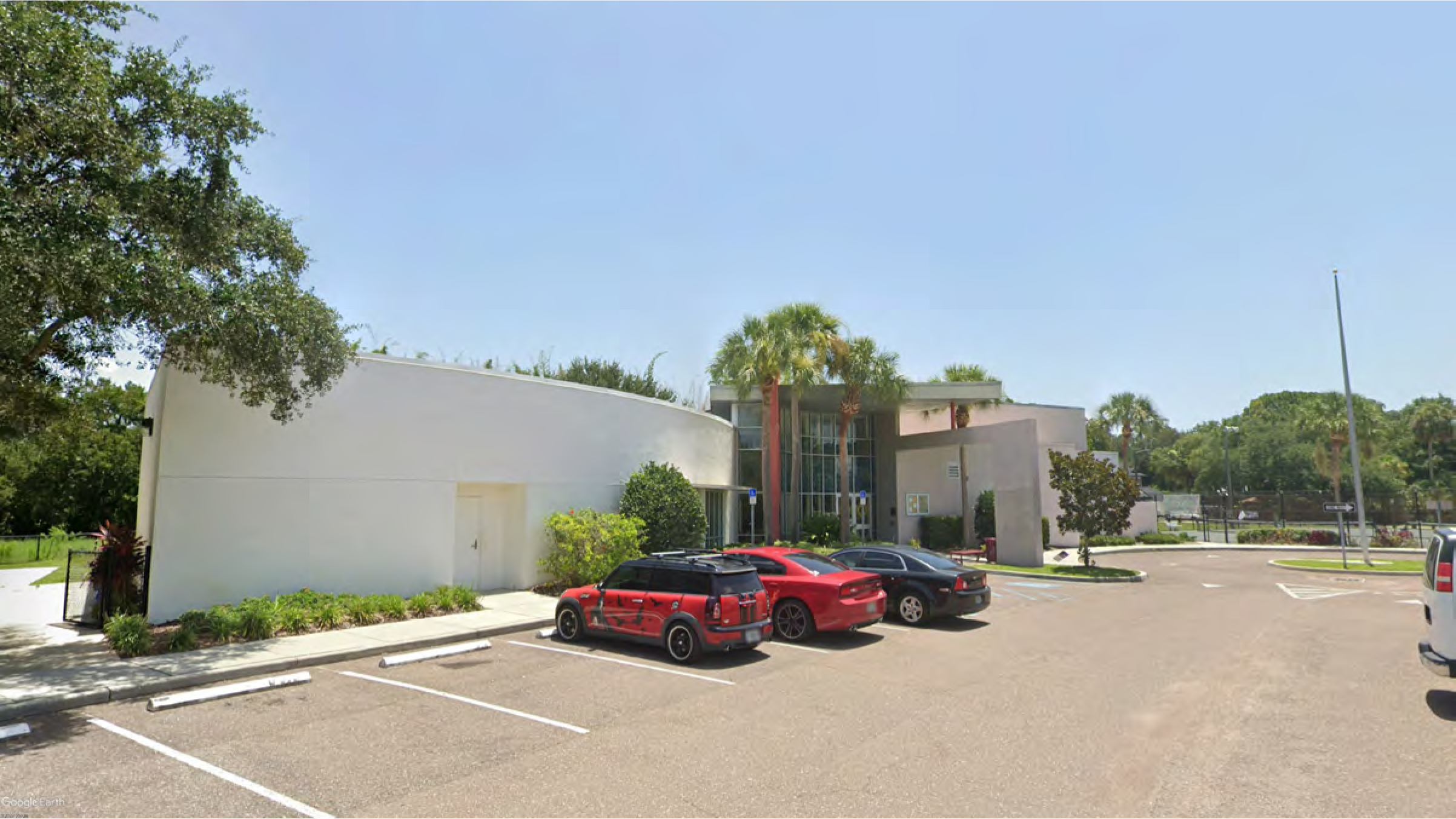 Modern commercial building with large windows and palm trees, featuring a parking lot with several cars.