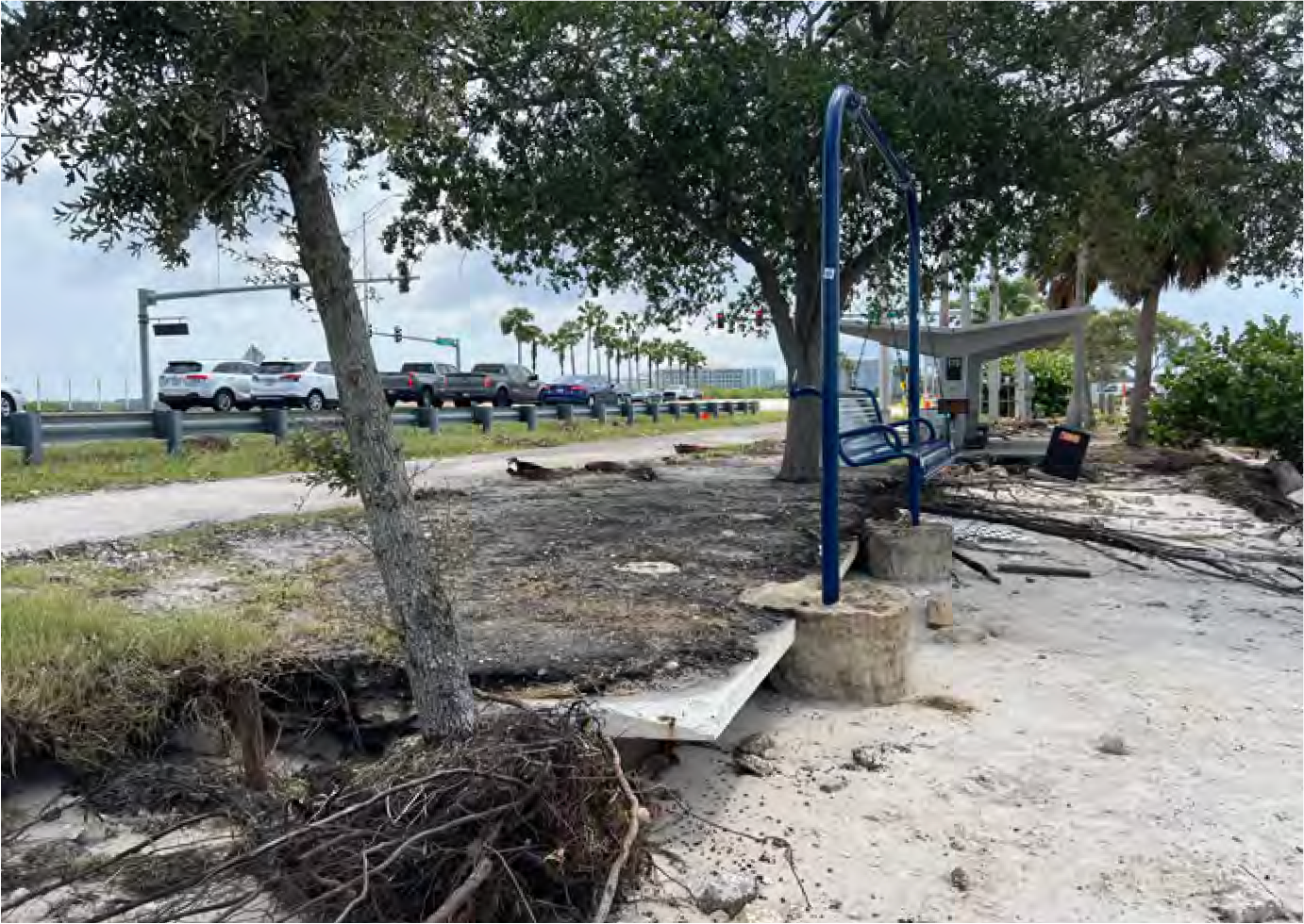 A partially eroded beach area with benches, trees, and nearby traffic visible in the background.