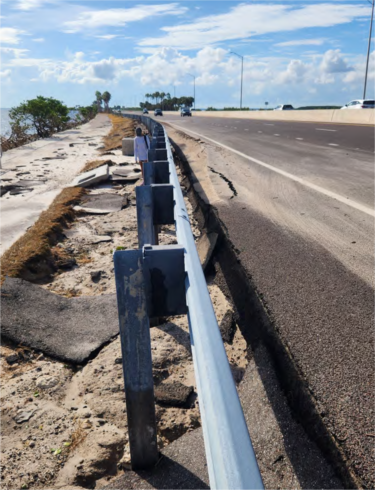 A damaged road shoulder with erosion near a highway, featuring a metal guardrail and a person walking along the shoreline.