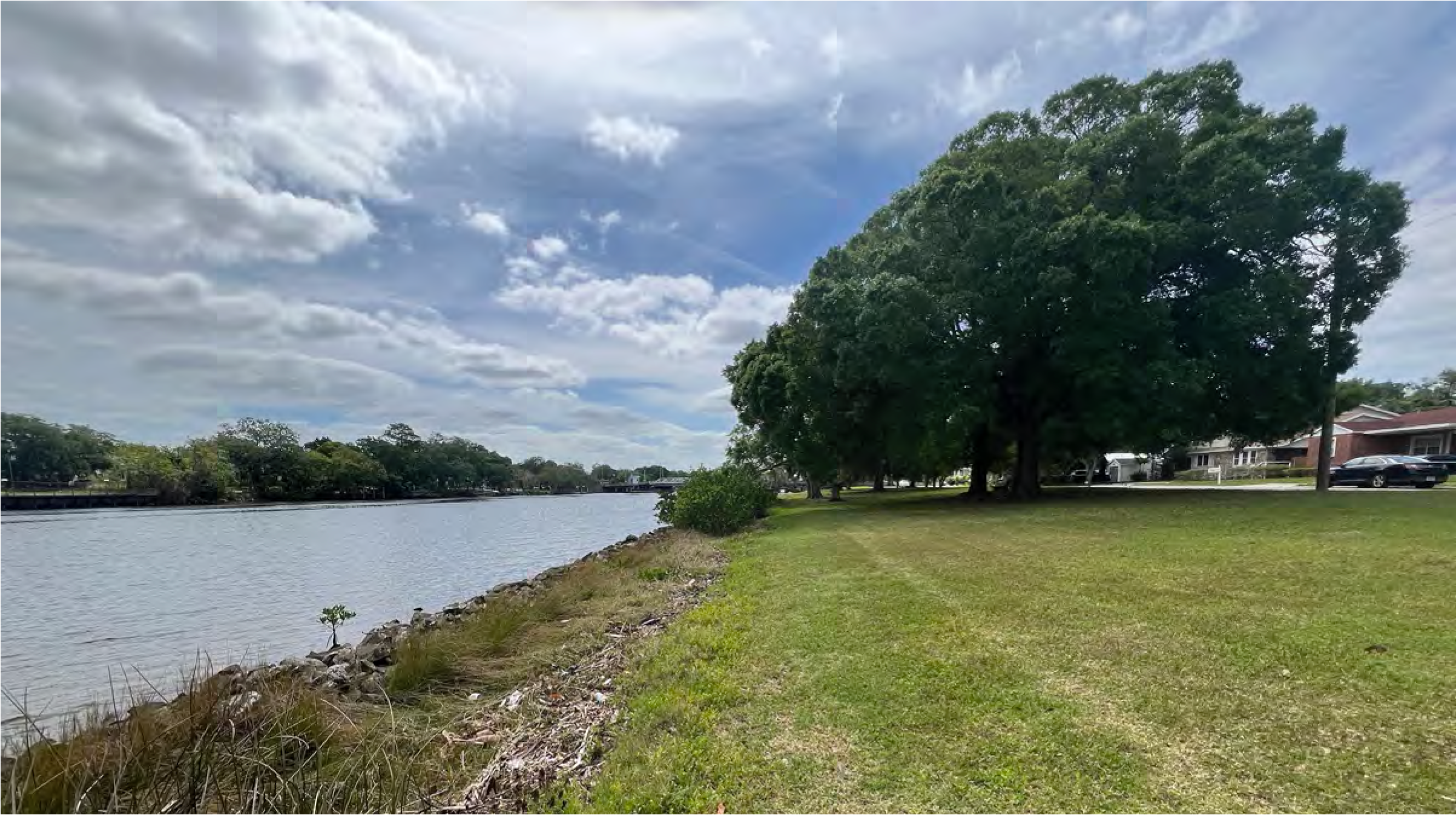 A scenic view of a riverbank with grassy land, trees, and cloudy blue sky.