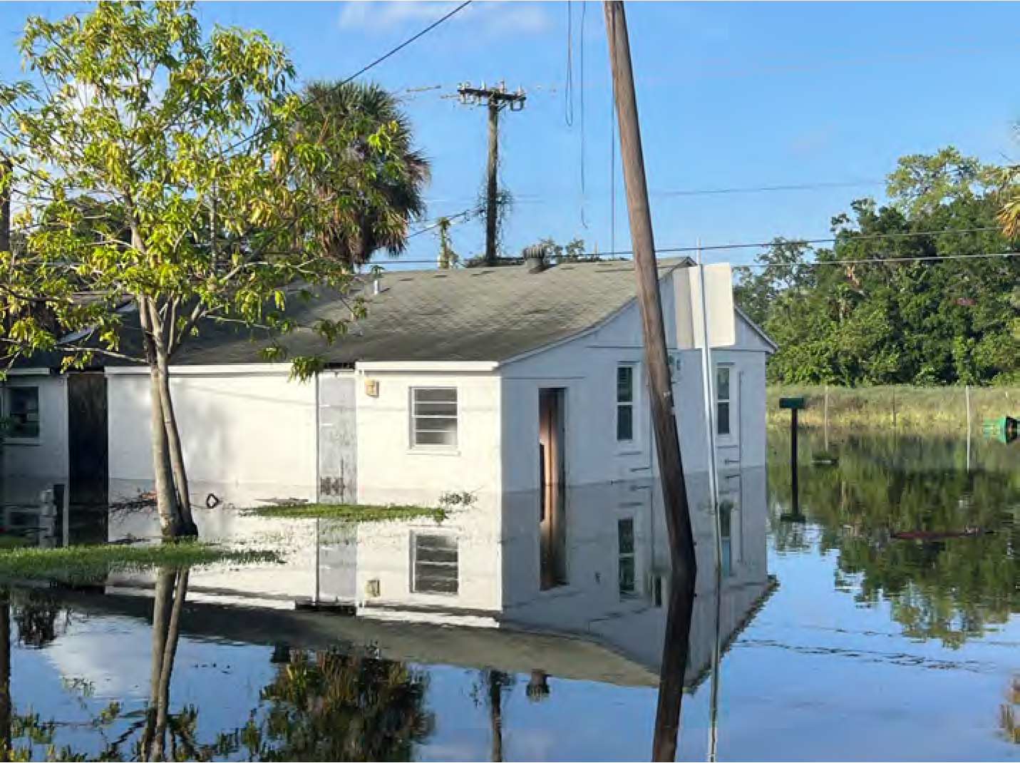 A flooded house reflected in water, with trees and utility poles visible in a clear blue sky.