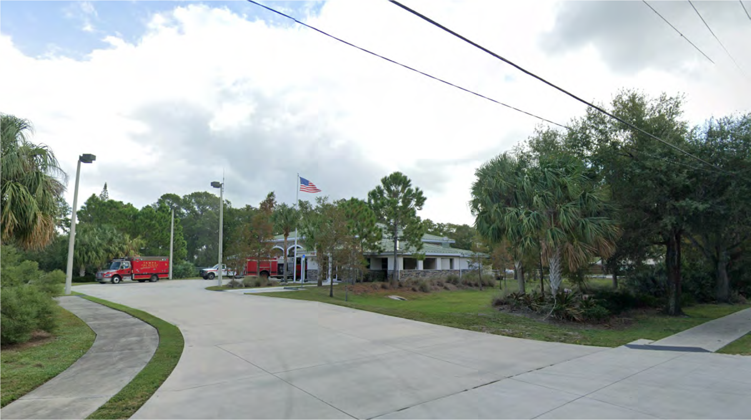 A view of a fire station with a large parking area, palm trees, and an American flag under a partly cloudy sky.