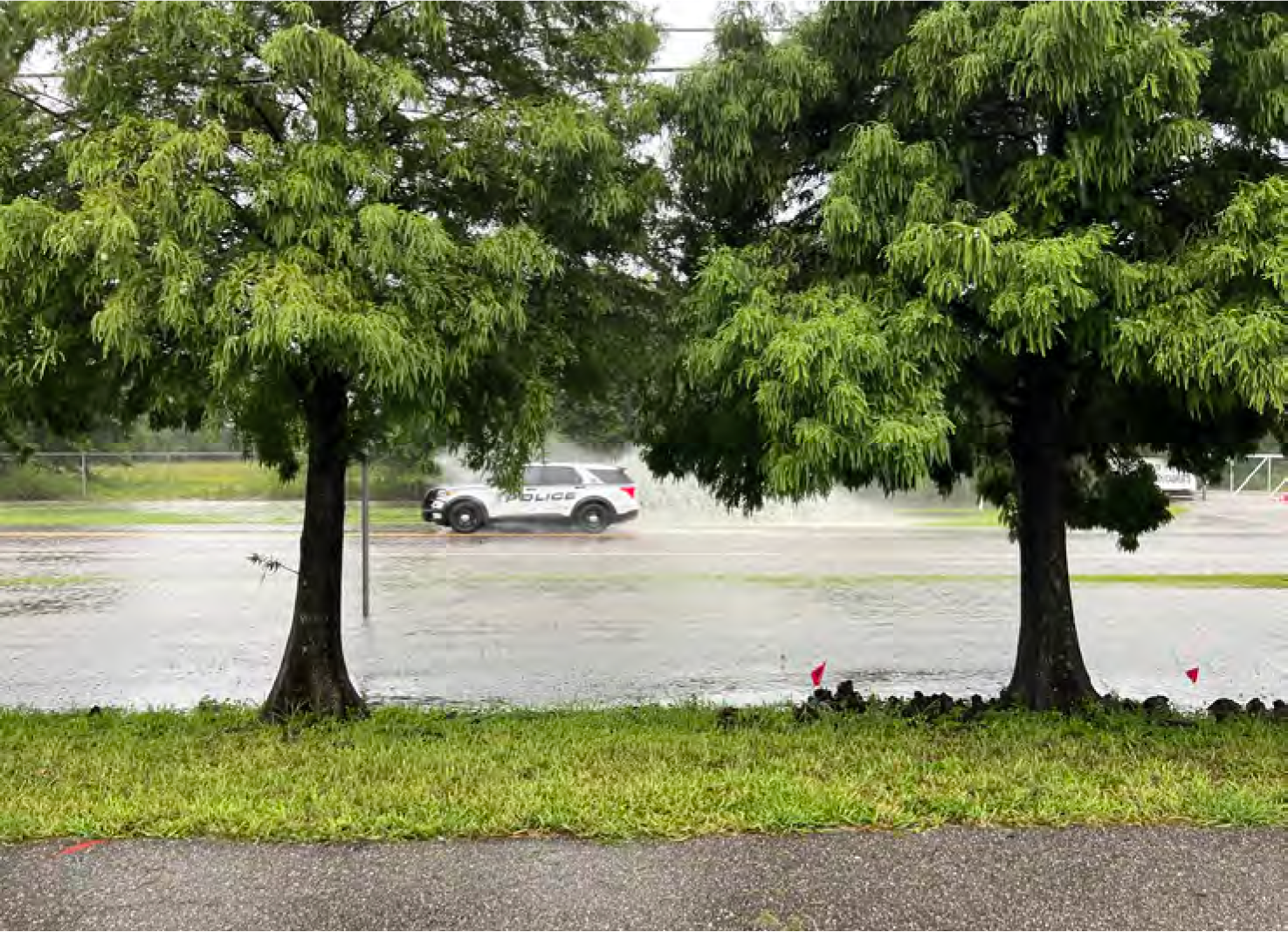 A police vehicle driving through a flooded street, surrounded by trees and grass.