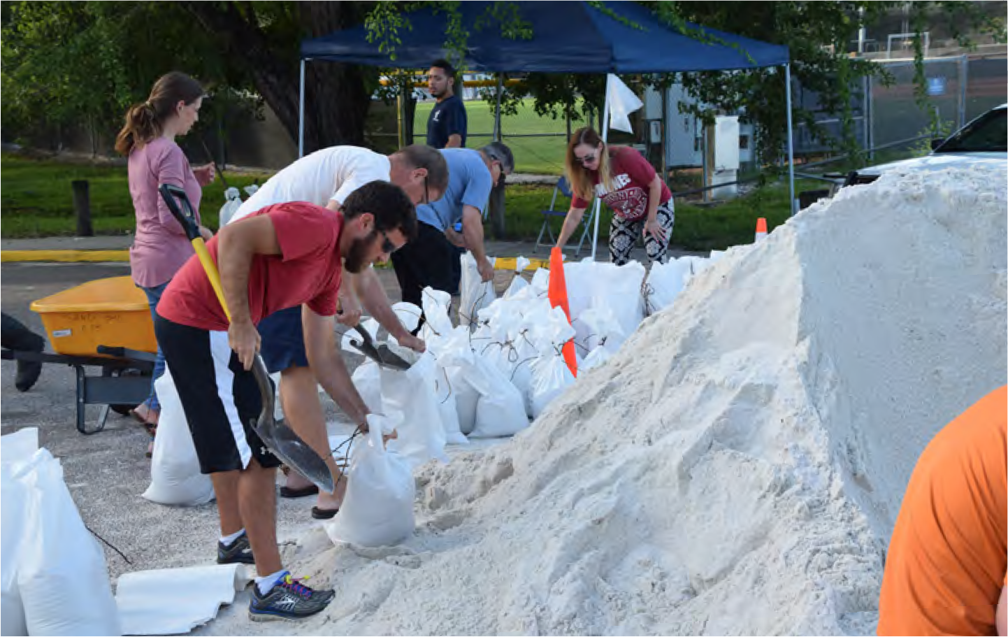 Volunteers filling sandbags at a community site in preparation for a flood, with tents and equipment in the background.