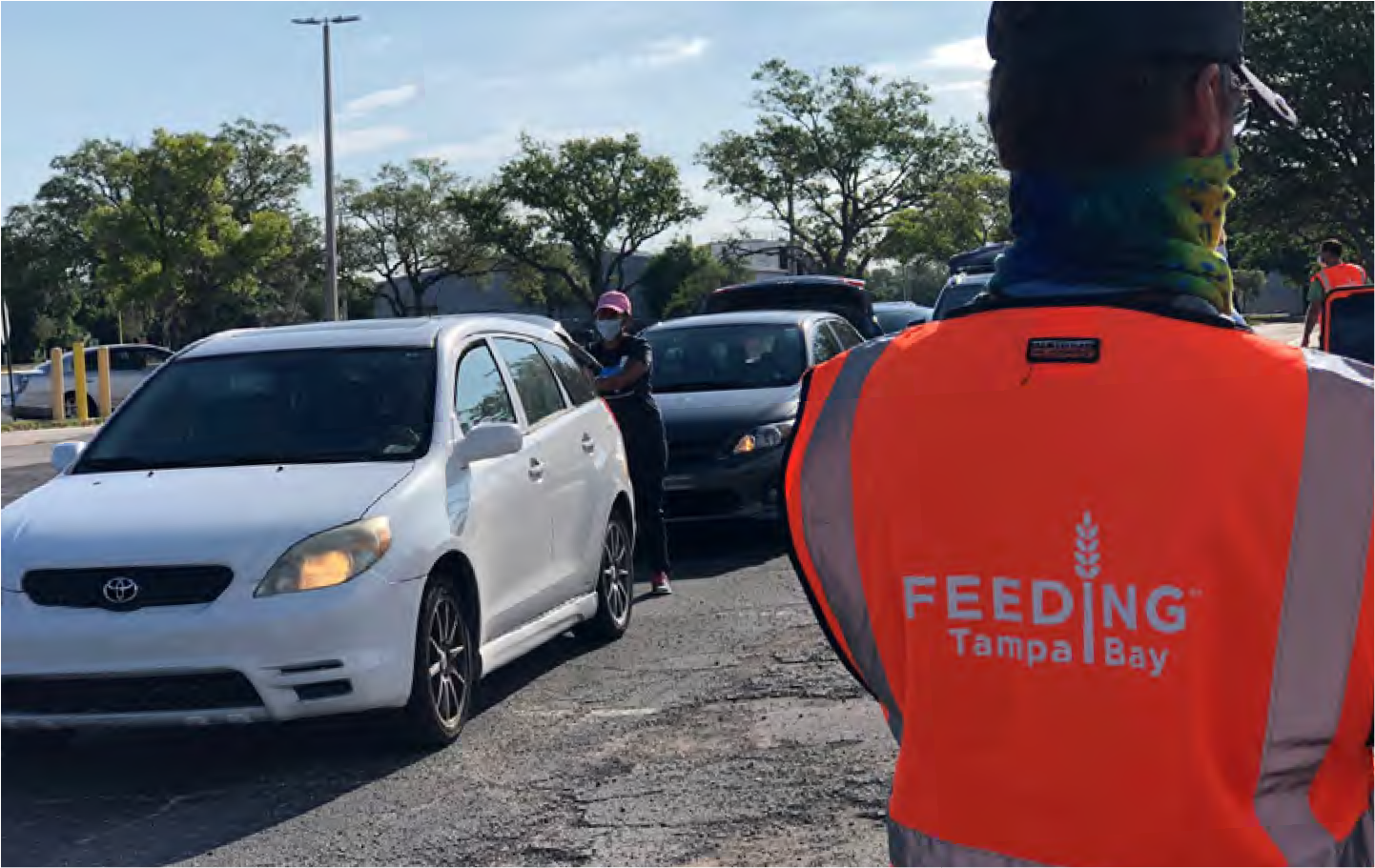 A volunteer in an orange vest assists cars in a food distribution event, with the logo 'Feeding Tampa Bay' visible.