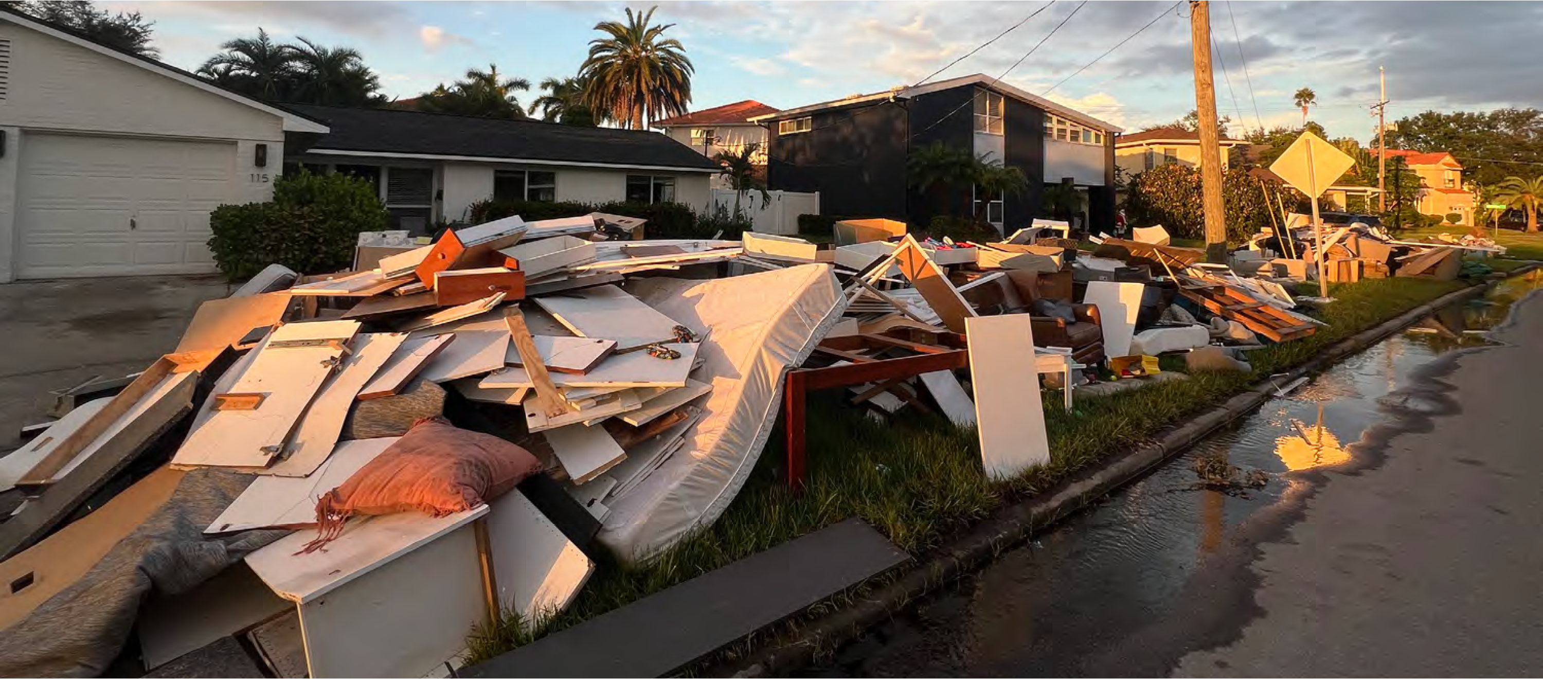 A large pile of discarded furniture and debris on a residential street, with palm trees and houses in the background.