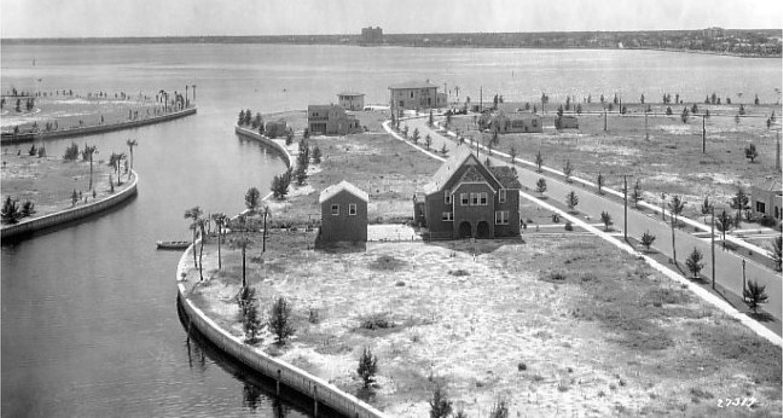 Aerial view of a waterway with houses and roads, surrounded by sparse land and palm trees, in a historical black and white photograph.