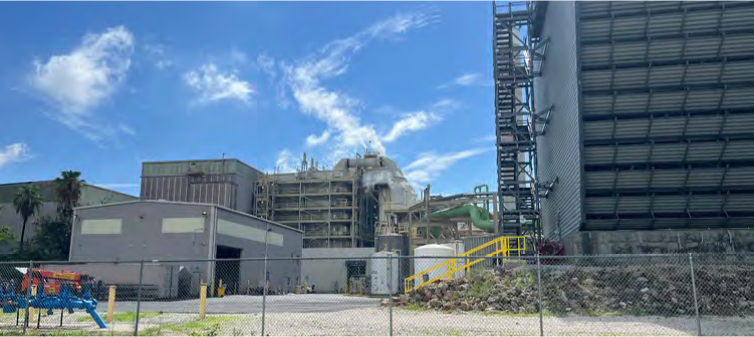 Industrial facility with large buildings, blue sky, and green pipes visible; fenced area in foreground.
