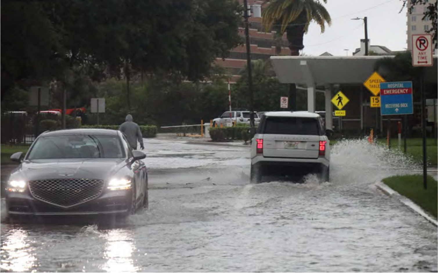 Two cars navigate a flooded street, with one vehicle creating splashes, and a pedestrian walking in the background.