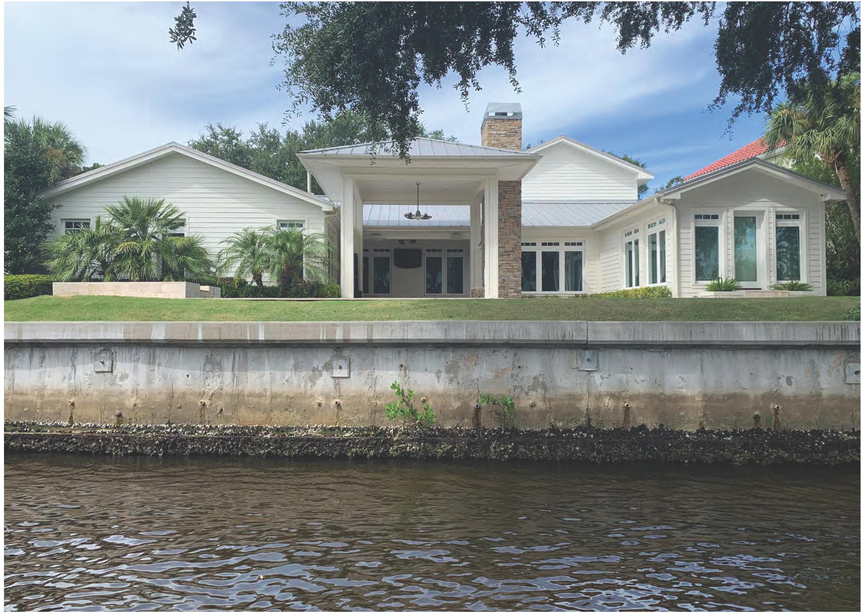 Waterfront view of a large, modern house surrounded by greenery with a stone accent and a flat roof.