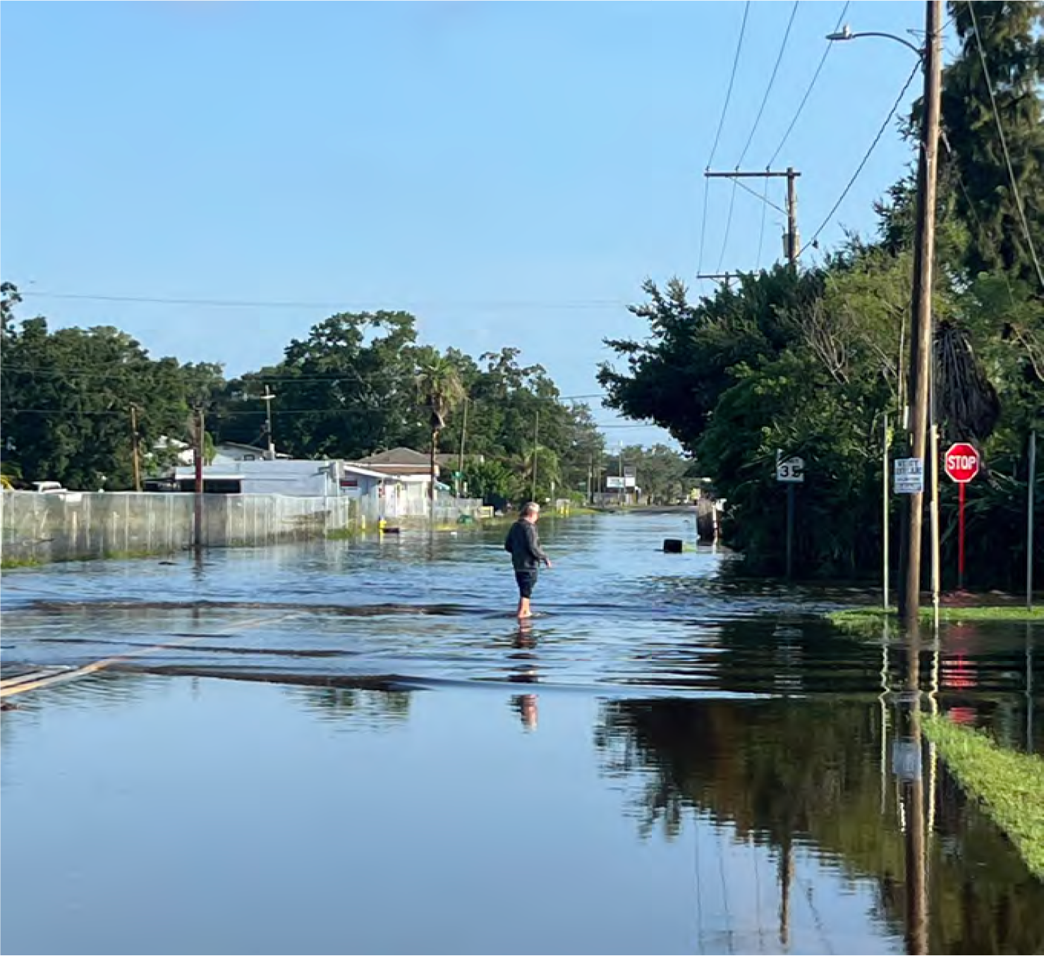 A person walking through a flooded street with standing water and power lines visible in the background.
