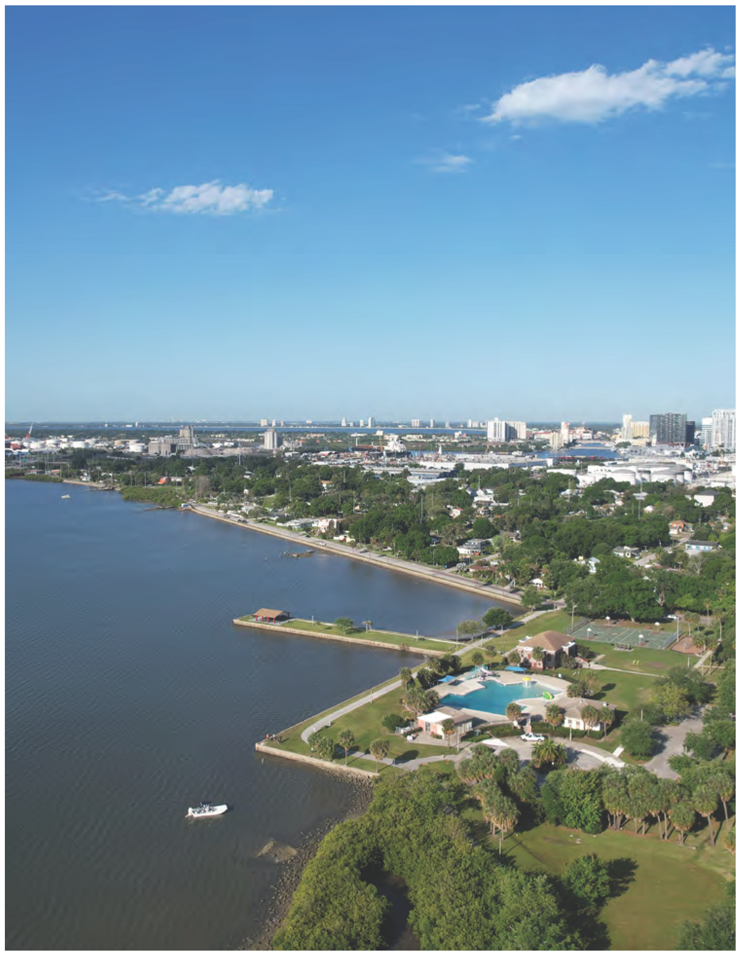 Aerial view of a waterfront area with lush greenery, buildings, and a pool, under a clear blue sky.