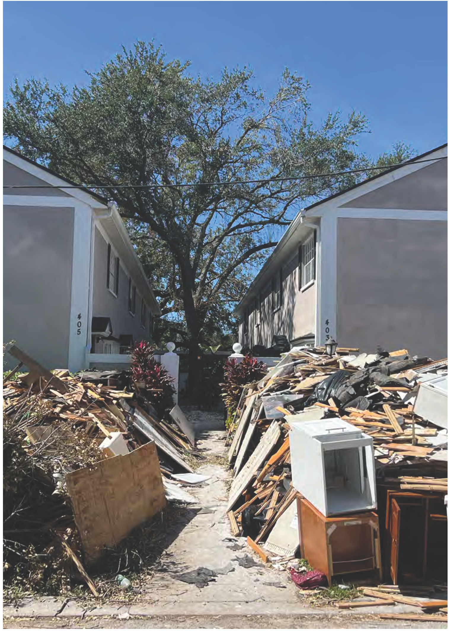 A pathway between two houses blocked by piles of debris and furniture under a clear blue sky.