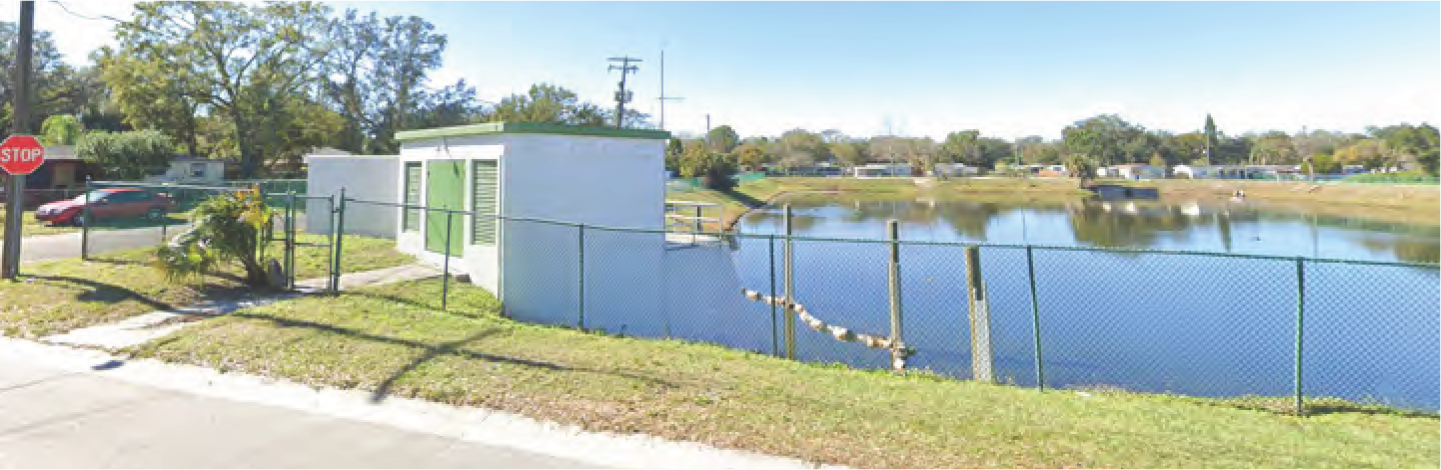 View of a pond surrounded by a fence, with a small building nearby and trees in the background.