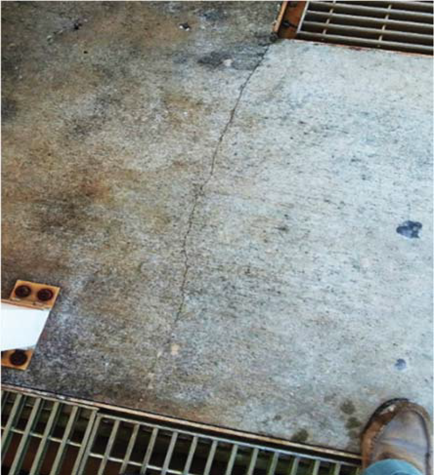 Close-up of a cracked concrete surface with a foot wearing a boot nearby, and metal grates in the foreground.