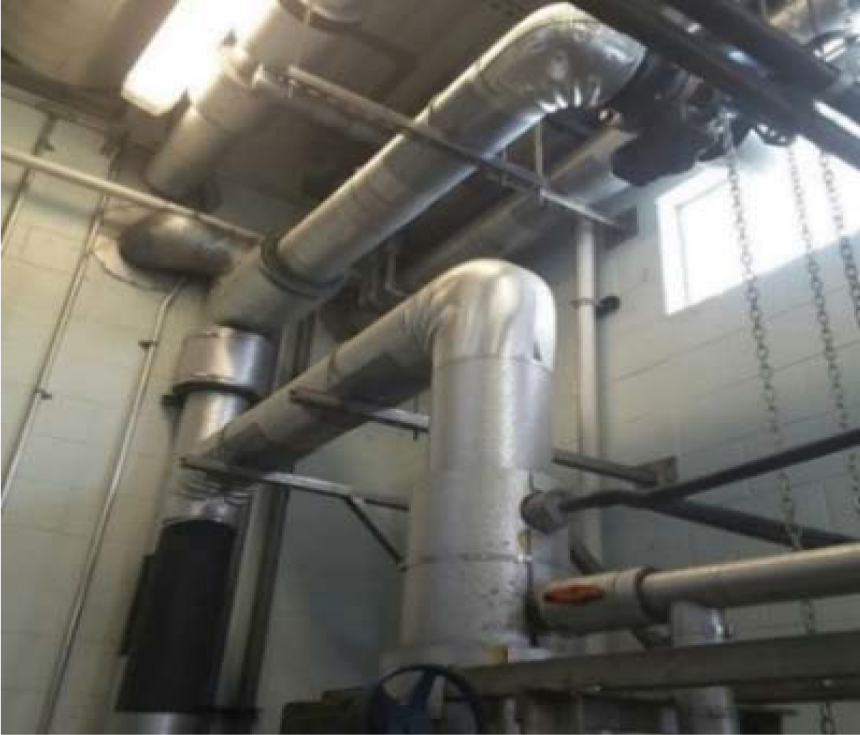 A network of industrial metal pipes and ducts connected to a wall, with visible joints and a window in the background.