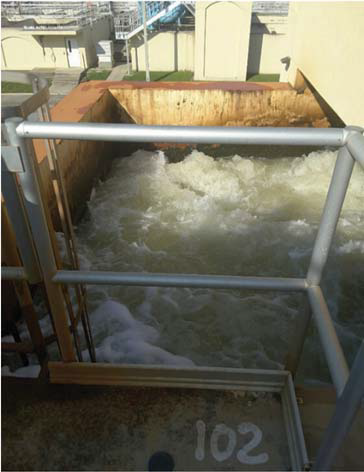 Water flowing through a facility, viewed from a metal railing with a partially visible pool area and surrounding structures.