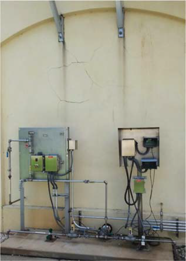 A wall-mounted control panel with green and black devices, pipes, and a visible crack in the wall behind it.