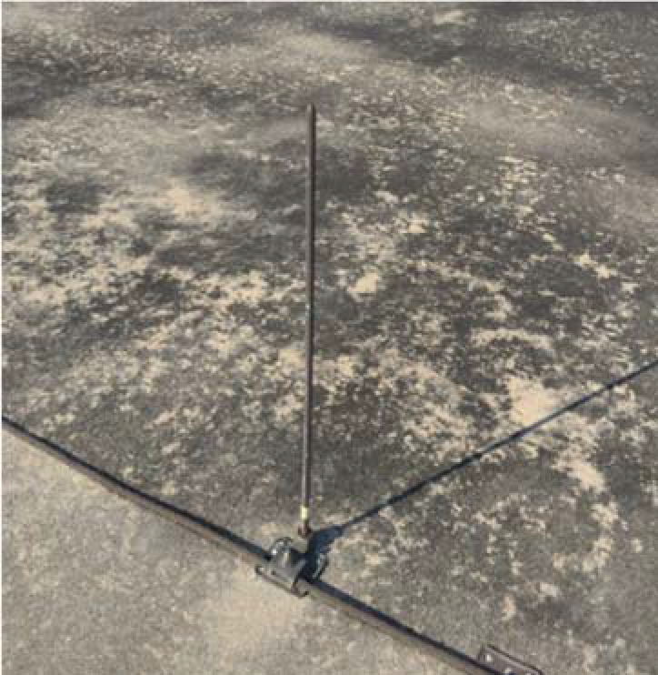 A vertical metal antenna mounted on a flat, textured surface with visible lines extending outward.