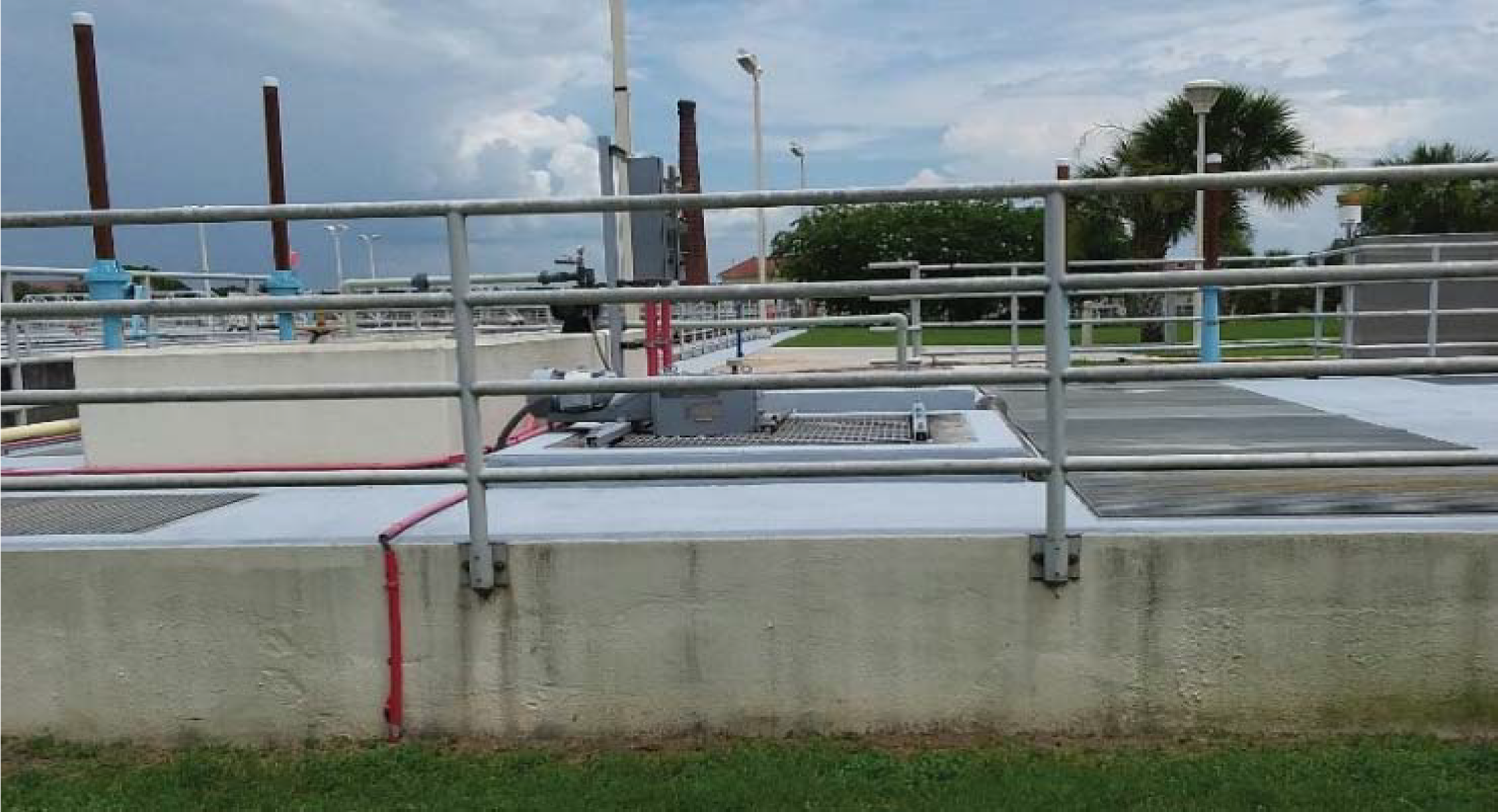 Infrastructure at a wastewater treatment facility, featuring railings, pipes, and equipment against a backdrop of grass and trees.