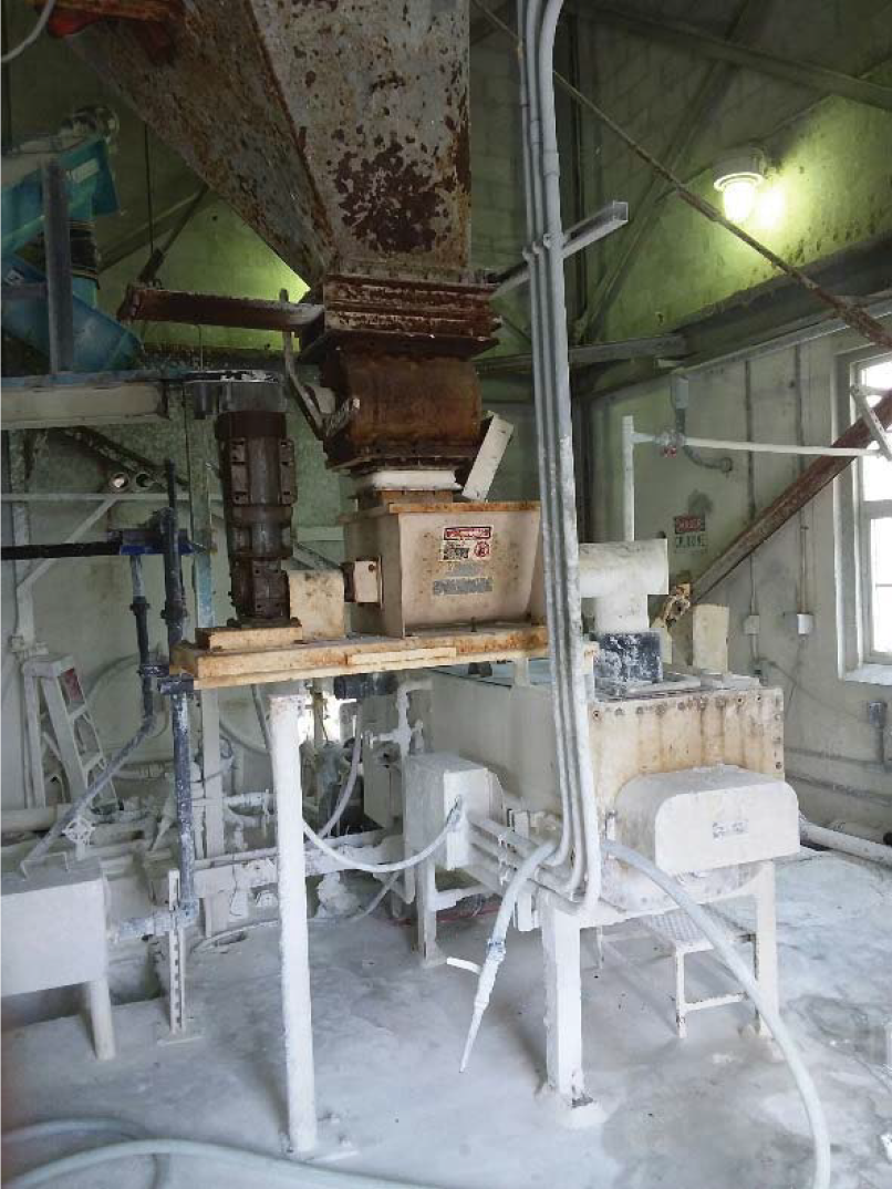 Industrial processing equipment in a factory, featuring a large hopper and machinery covered in white powder, set in a dimly lit environment.