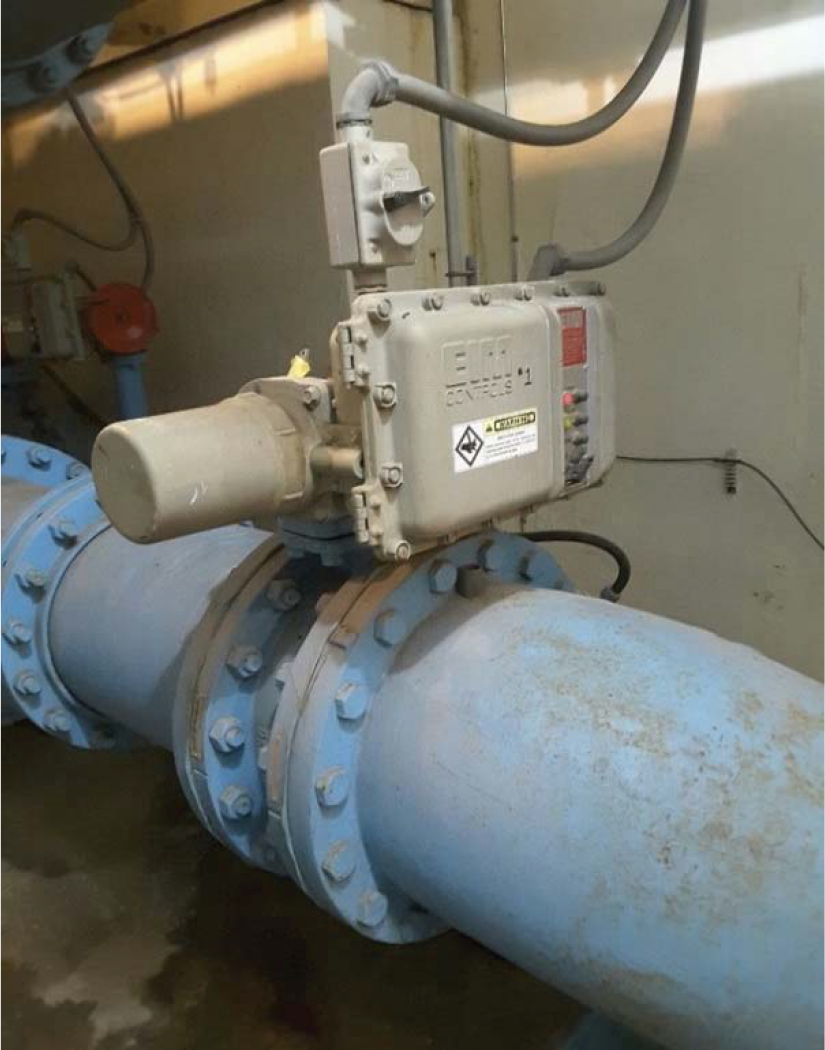 A large blue pipe with a control valve and electrical components attached, located indoors in an industrial setting.