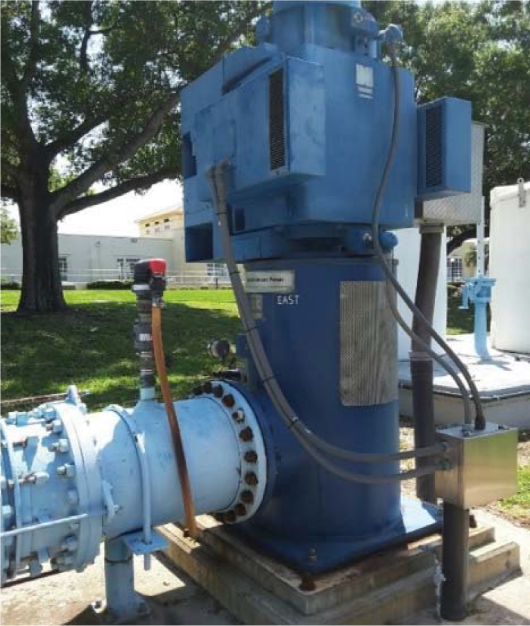 Industrial blue pump with connected piping, situated outdoors near trees and buildings.