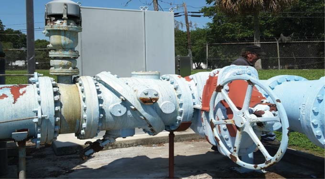A large rusted water valve and pipes with a person operating a valve in the background on a sunny day.