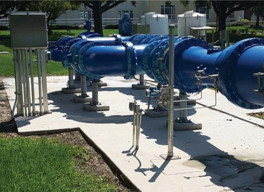 Blue industrial piping system on a concrete slab, surrounded by grass and trees.