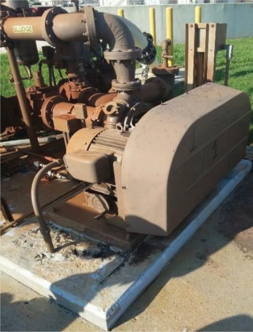 An industrial pump system with rusted pipes and machinery, situated on a concrete base in a grassy area.