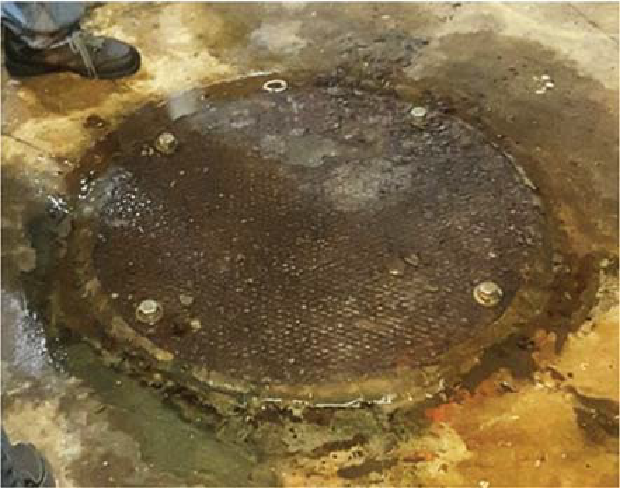 A large, rusty manhole cover surrounded by water on a concrete floor, with a person's feet visible nearby.
