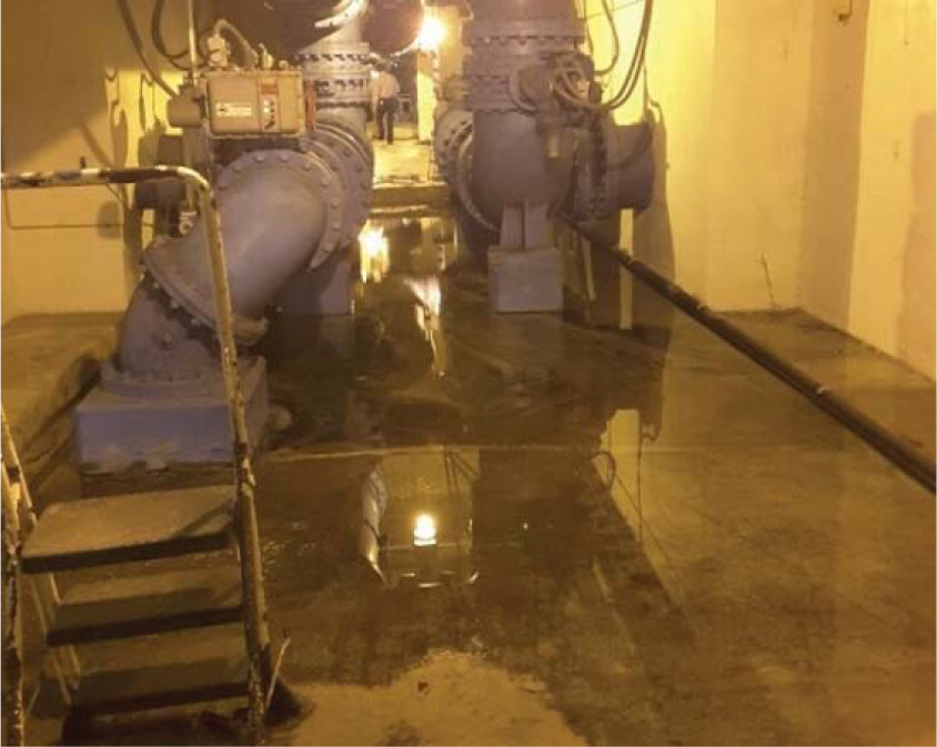 Industrial pipes in a dimly lit basement with water pooling on the concrete floor.